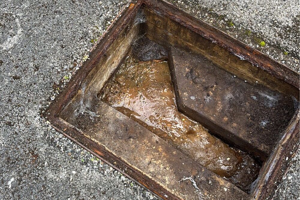Open outdoor drainage chamber with standing water and debris inside, surrounded by concrete paving.